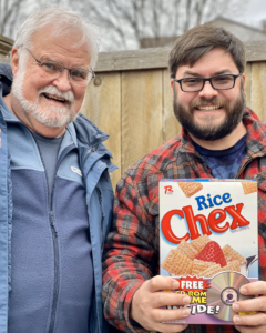 A father and his adult son posing with a box of Rice Chex cereal.